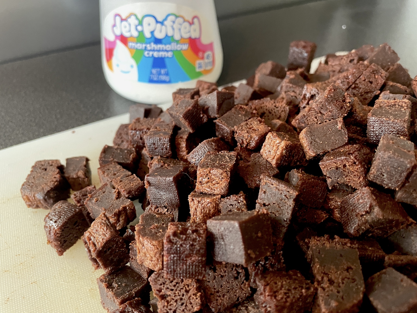 a cutting board with gooey brownie bits for ice cream chopped up and a jar of marshmallow fluff in the background